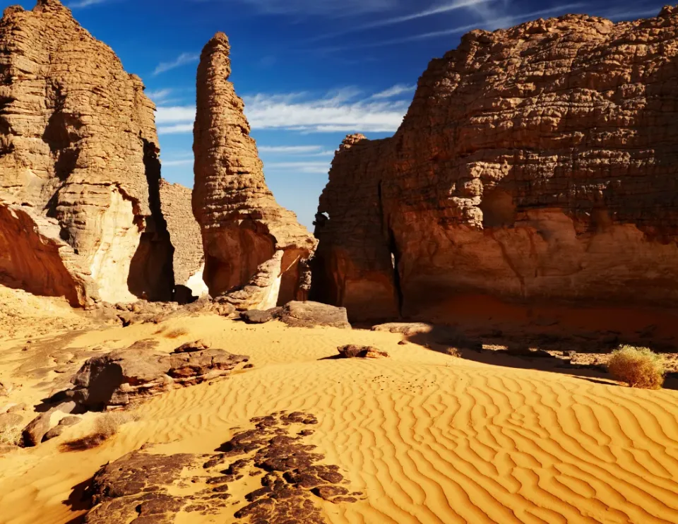 Tall sandstone rock formations rise from golden rippled desert sand under a clear, bright blue sky.