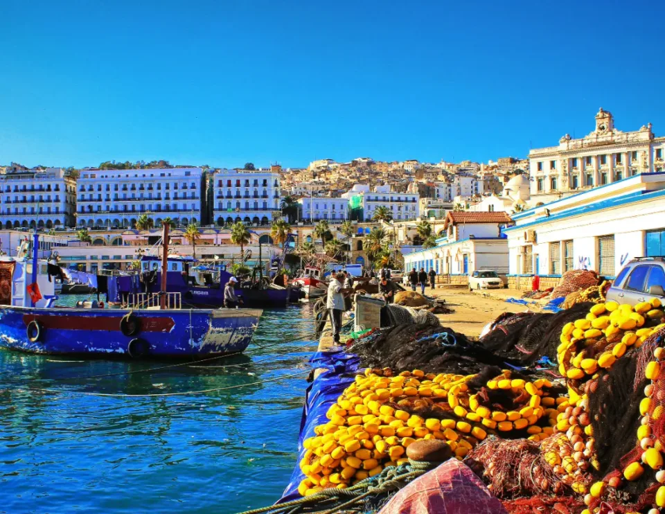 A blue fishing boat at a harbor quay with piles of yellow fishing nets in the foreground and city buildings behind.