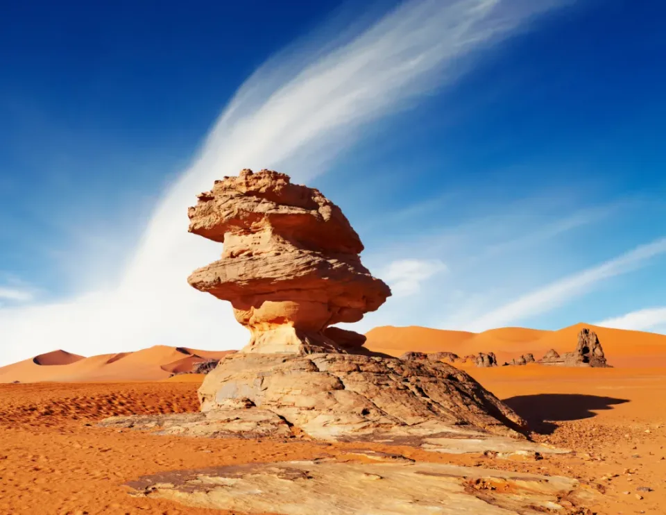 A large, weathered sandstone rock formation stands in the middle of a vast, orange desert under a bright blue sky.